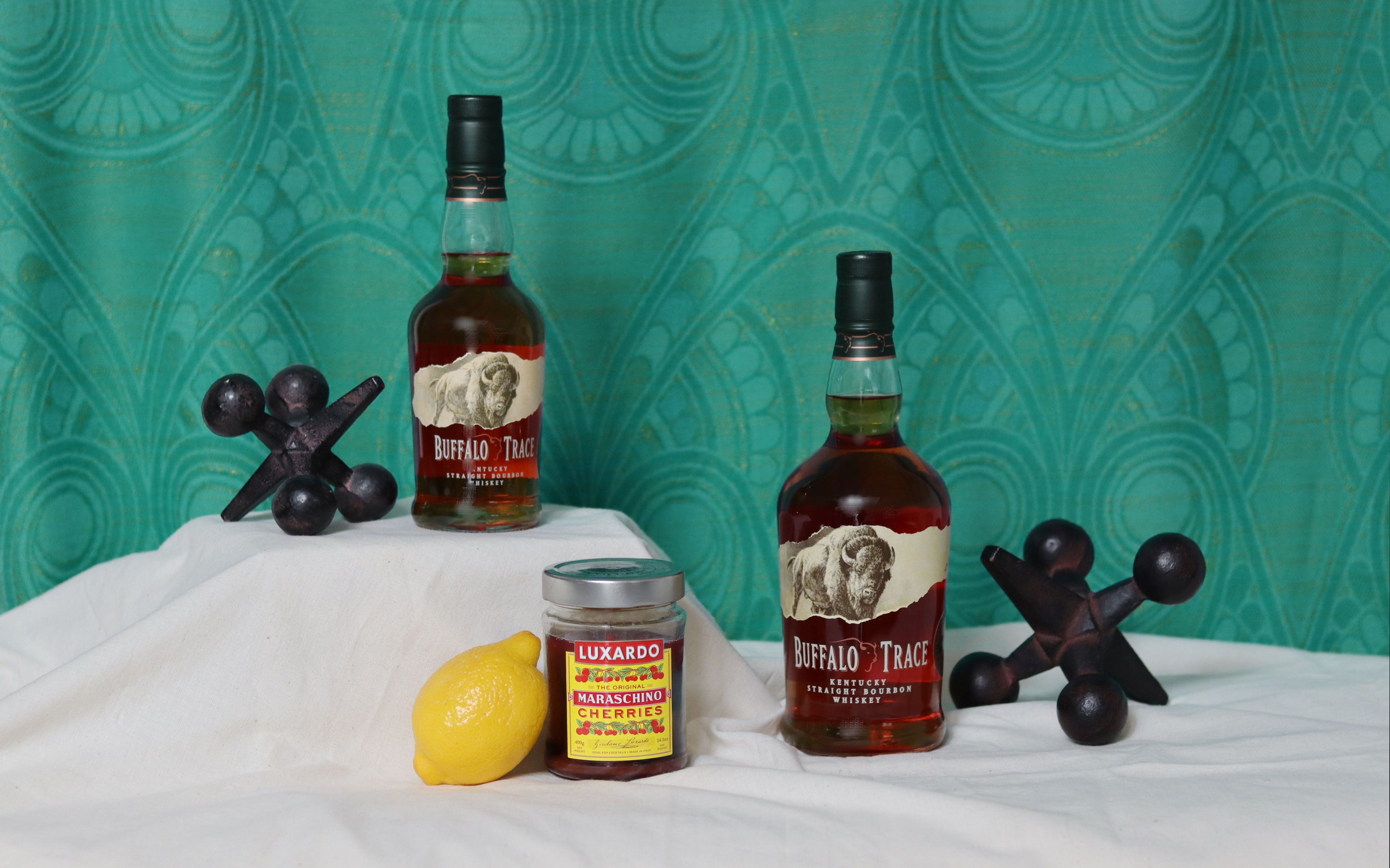 Two bottles of Buffalo Trace bourbon with a lemon, jar, and decorative items on a white surface against a teal patterned wall.