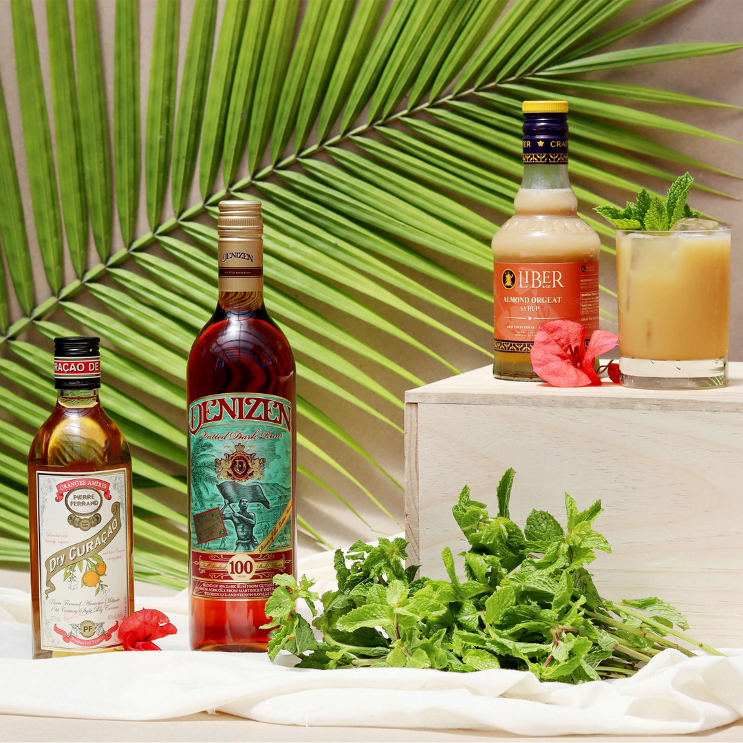 Three bottles of alcohol on a table with a tropical leaf and cocktail in the background