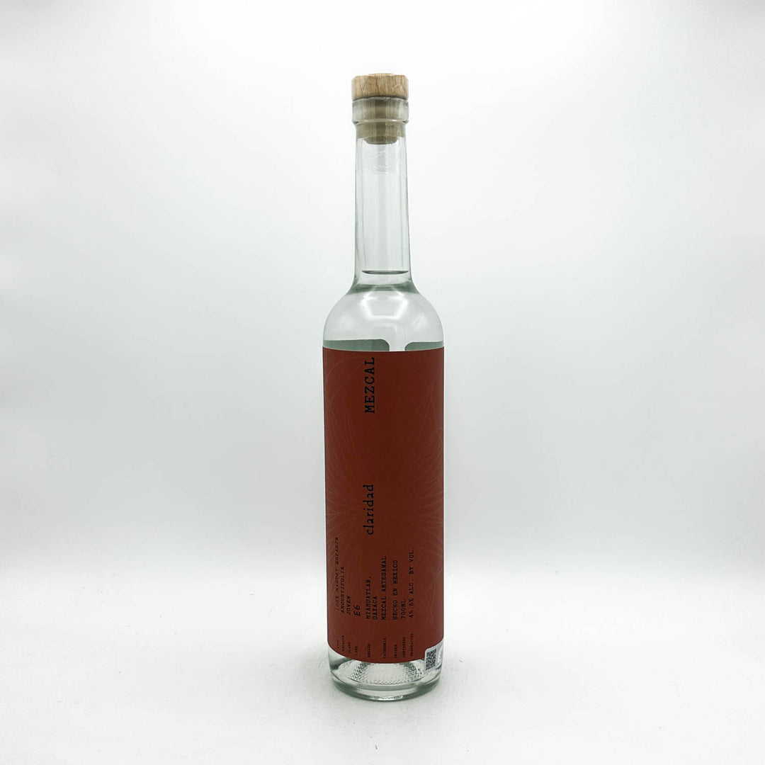 A clear glass bottle with a cork stopper and a vertical reddish-brown label, photographed against a plain light gray studio background.