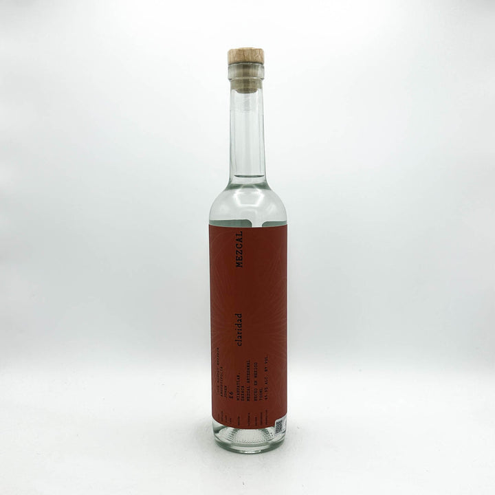 A clear glass bottle with a cork stopper and a vertical reddish-brown label, photographed against a plain light gray studio background.