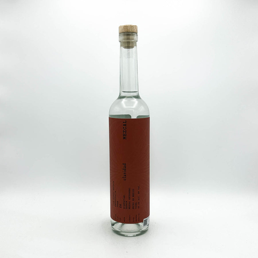 A clear glass bottle with a cork stopper and a vertical reddish-brown label, photographed against a plain light gray studio background.