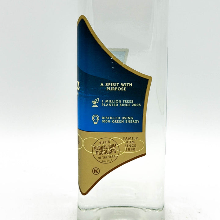 A clear glass bottle with a blue and gold label, photographed against a plain white background. The label features text including 'A SPIRIT WITH PURPOSE' and '1 MILLION TREES PLANTED SINCE 2005'.