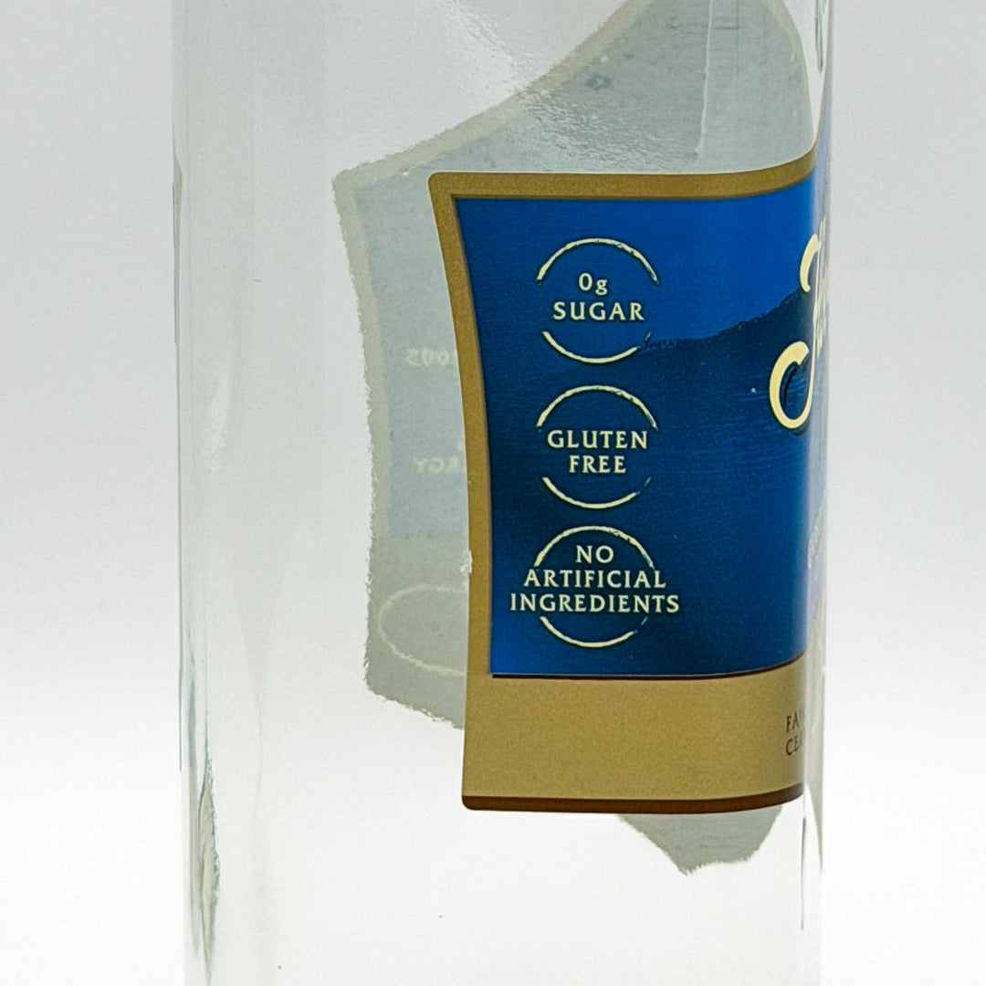 Close-up of a clear glass bottle with a blue and gold label featuring text that reads '0g SUGAR', 'GLUTEN FREE', and 'NO ARTIFICIAL INGREDIENTS'.