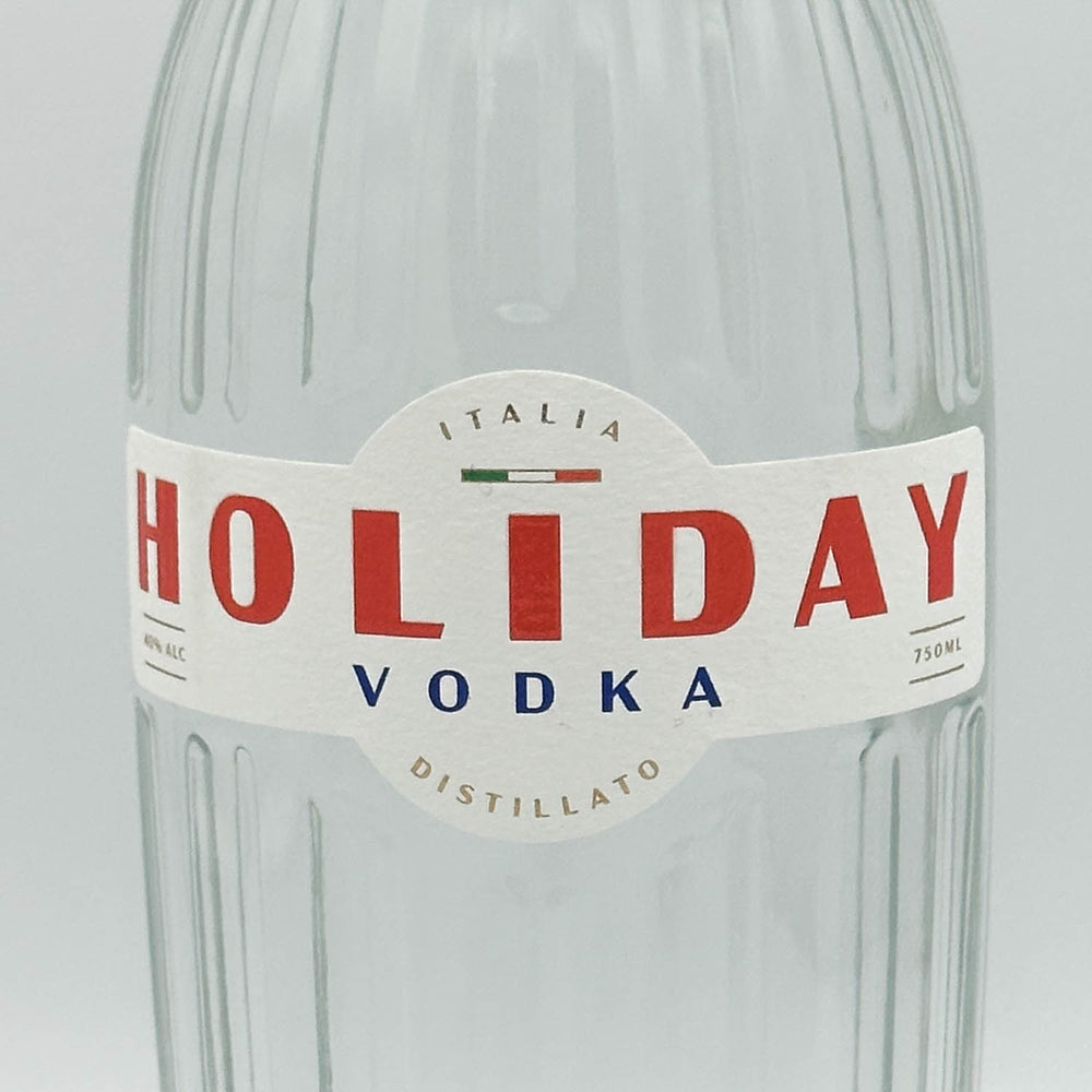 A close-up of a clear ribbed glass bottle with a white paper label reading HOLIDAY VODKA, with large red lettering and smaller blue text, on a light gray background.