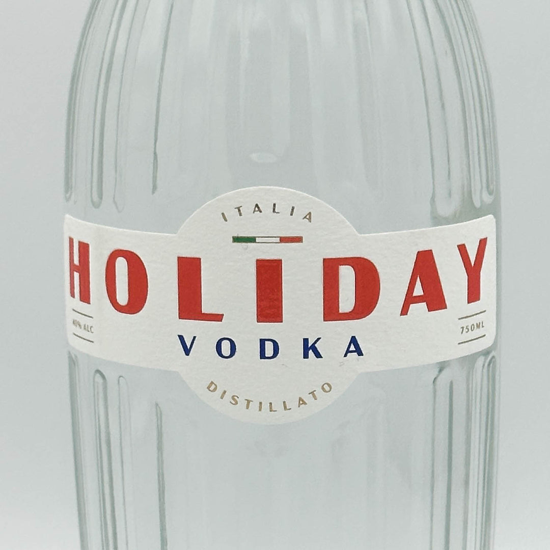 A close-up of a clear ribbed glass bottle with a white paper label reading HOLIDAY VODKA, with large red lettering and smaller blue text, on a light gray background.