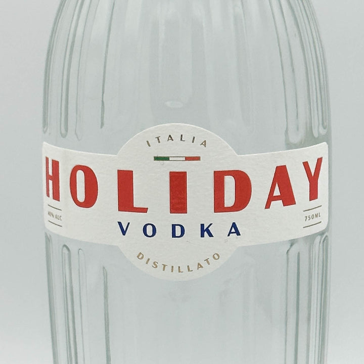 A close-up of a clear ribbed glass bottle with a white paper label reading HOLIDAY VODKA, with large red lettering and smaller blue text, on a light gray background.