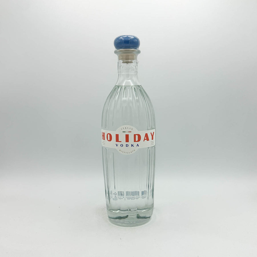 A clear glass bottle of Holiday Vodka with a ribbed texture, a white label with red lettering, and a blue cork stopper, set against a plain light gray background.