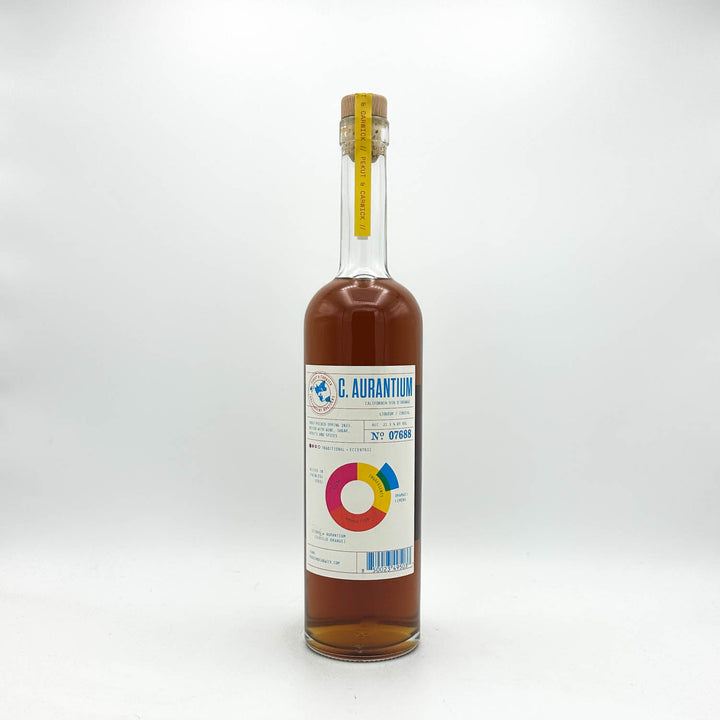 A clear glass bottle of C. Aurantium syrup with amber-brown liquid inside, a white label with blue and multicolor graphics, and a cork stopper with a yellow seal, photographed on a light gray background.