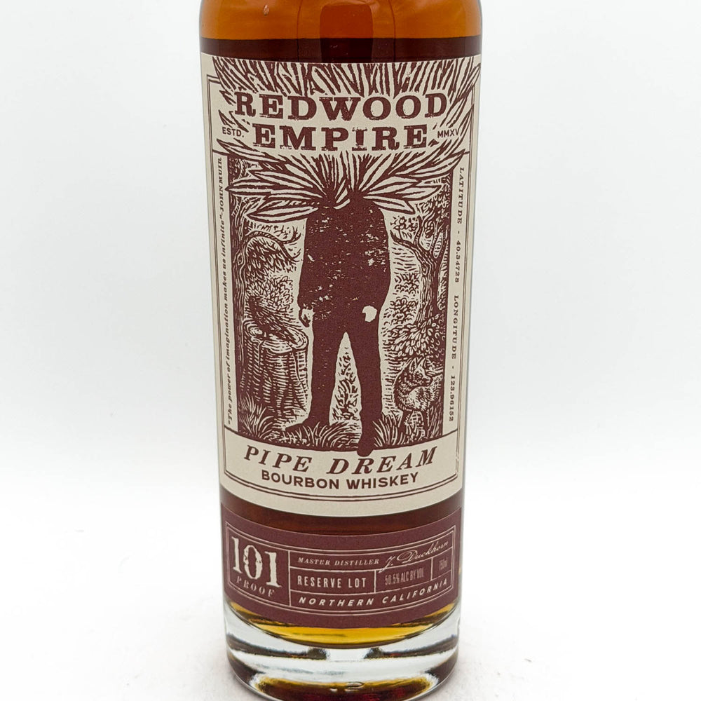 A bottle of Redwood Empire Pipe Dream Bourbon Whiskey, showing the amber liquid and a detailed cream and dark red label with an illustration of a figure.