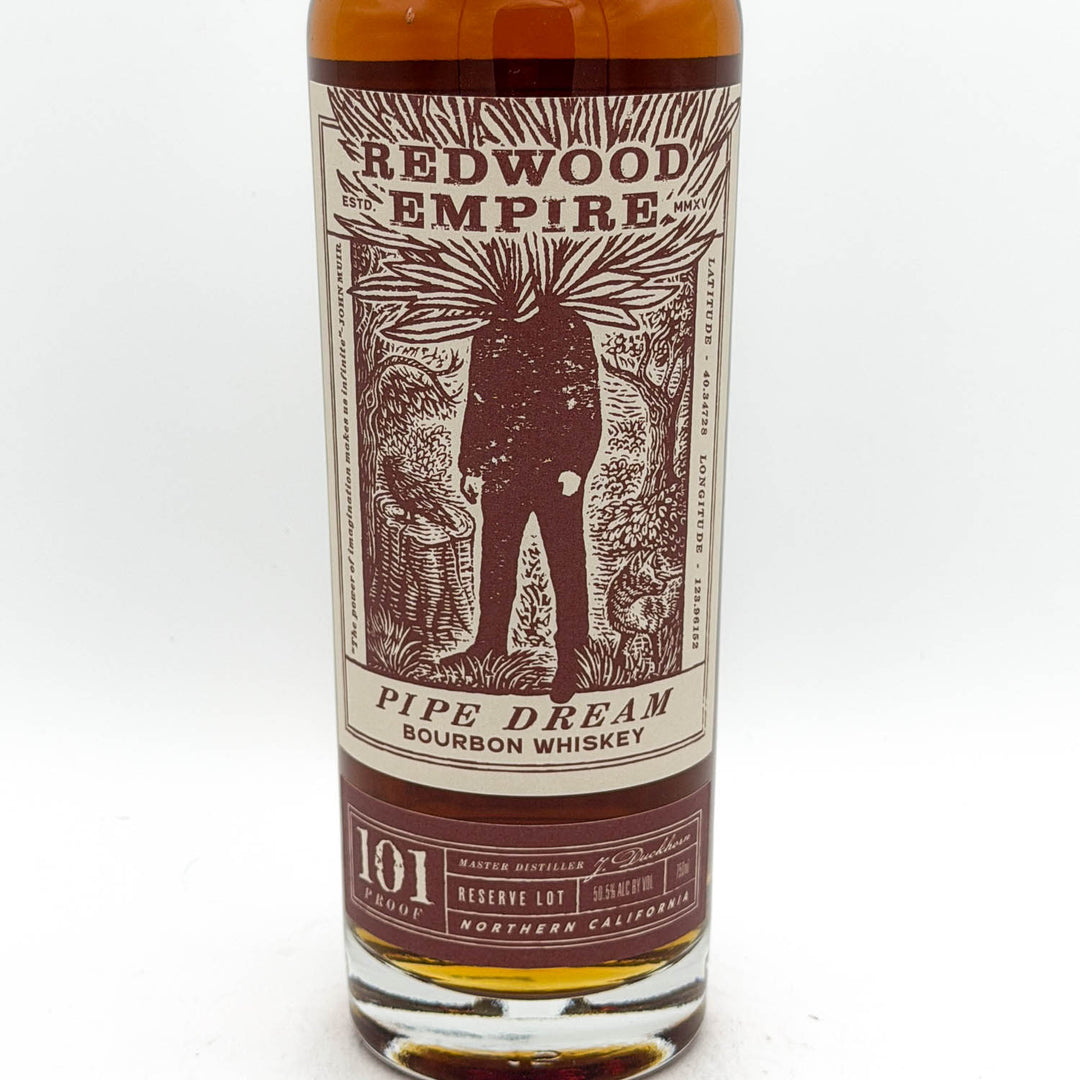 A bottle of Redwood Empire Pipe Dream Bourbon Whiskey, showing the amber liquid and a detailed cream and dark red label with an illustration of a figure.