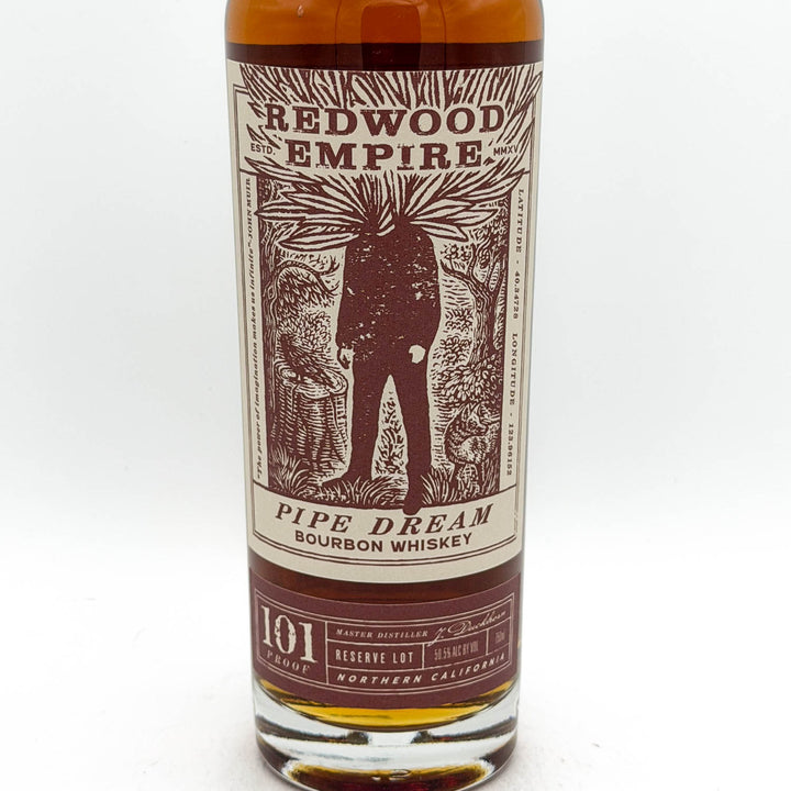 A bottle of Redwood Empire Pipe Dream Bourbon Whiskey, showing the amber liquid and a detailed cream and dark red label with an illustration of a figure.